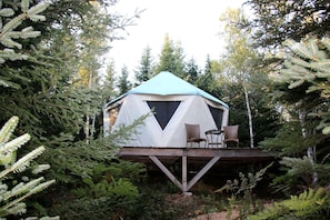 Una cúpula geodèsica, una tenda amb plataforma i cadires, envoltada d'un bosc dens.