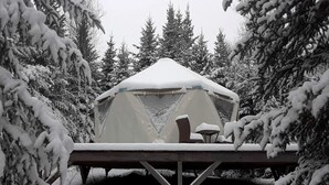 A snow-covered tent with a table and chairs, surrounded by snow-laden trees.