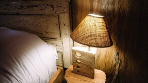 A wooden bedside table with a woven lamp, a bed with white bedding, and a rustic wall. - Luxurious Woodland Cabin in Nova Scotia (Baddeck)