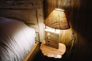 Une table de chevet en bois surmontée d’une lampe au design tressé, un lit avec une literie blanche et un mur au style rustique.