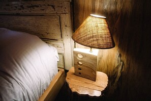 A wooden bedside table with a woven lamp, a bed with white bedding, and a rustic wall. - Luxurious Woodland Cabin in Nova Scotia (Baddeck)