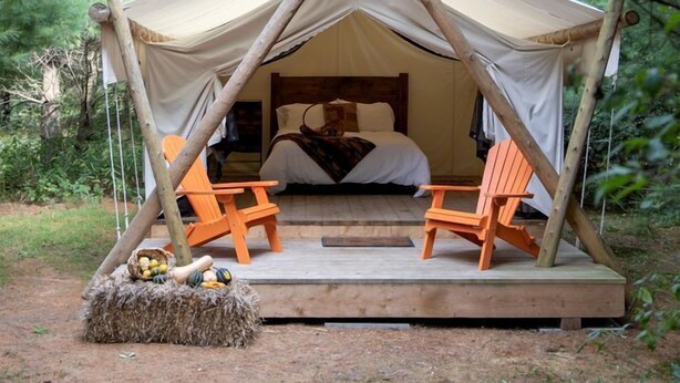 A tent with a bed, two chairs, and a hay bale with vegetables.