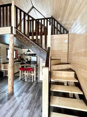 Un escalier en bois avec une rampe, un coin repas avec des chaises rouges et une cuisine en arrière-plan.
