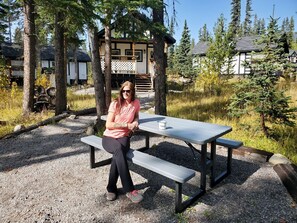 Una persona sentada en una mesa de picnic en un área boscosa, con una cabaña al fondo.