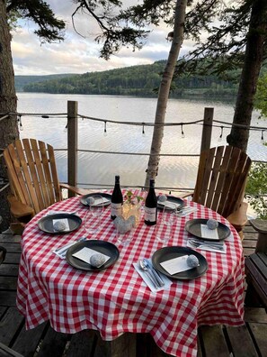 A table set outdoors with a checkered tablecloth, plates, glasses, and bottles, surrounded by wooden chairs. - Cardinal's Inn Cabin Rental in Nova Scotia (Baddeck)