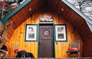 A wooden cabin with a dark door, two windows with white frames, and a sign above the door.