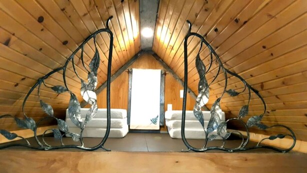 Una habitación de madera en el ático con techo inclinado, baranda decorativa de metal y vista a un sofá blanco a través de un umbral.