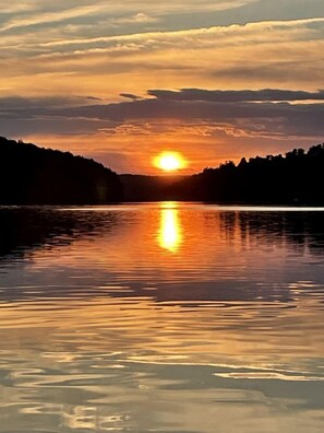 A calm lake at sunset with a reflection of the sun and a silhouette of trees on the opposite shore.