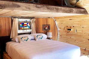 Ein Schlafzimmer einer Holzhütte mit Bett, Vorhängen, einem Buntglasfenster und einer Wandleuchte.