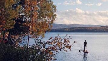 Eine Person steht auf einem Paddleboard auf einem ruhigen See, im Hintergrund sind Bäume am Ufer und Berge zu sehen.