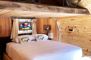 A wooden cabin bedroom with a bed, curtains, a stained glass window, and a wall-mounted light fixture.
