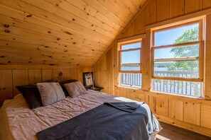 A wooden cabin bedroom with a bed, pillows, a nightstand, a lamp, and large windows overlooking a lake.