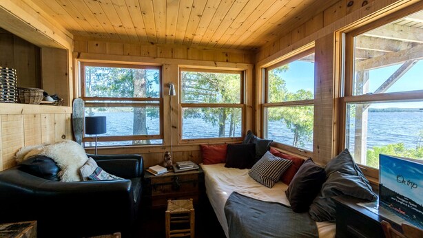 Ein Innenraum einer Hütte mit einem Bett, zwei Stühlen, einem kleinen Tisch und großen Fenstern mit Blick auf einen See.