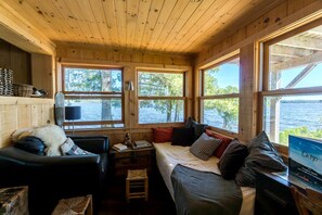 A cabin interior with a bed, two chairs, a small table, and large windows overlooking a lake.