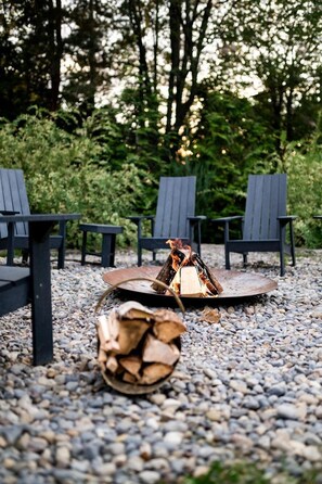 A fire pit with logs, surrounded by Adirondack chairs on a gravel surface. - Lakefront Chalet in Prince Edward County (Prince Edward County)