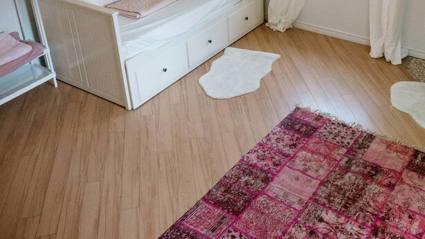 Ein Zimmer mit einem weißen Bett, einem Deckenventilator, einem Fenster mit weißen Vorhängen und einem pinkfarbenen Teppich mit Muster.