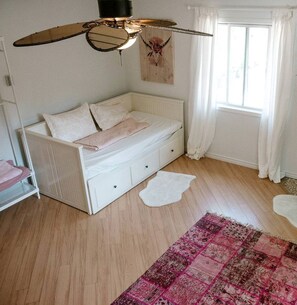 A room with a white bed, a ceiling fan, a window with white curtains, and a pink patterned rug. - Lakefront Chalet in Prince Edward County (Prince Edward County)