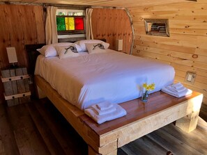 A wooden cabin-style bedroom with a bed, pillows, a nightstand, a lamp, and a small window with curtains. - Herons Haven Cabin in Nova Scotia (Baddeck)