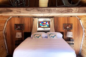 A cozy wooden cabin bedroom with a bed, curtains, and a window with a colorful view. - Herons Haven Cabin in Nova Scotia (Baddeck)