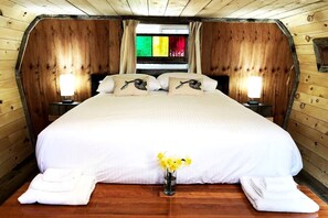 A wooden cabin-style bedroom with a large bed, two bedside lamps, and a window with colorful curtains. - Herons Haven Cabin in Nova Scotia (Baddeck)