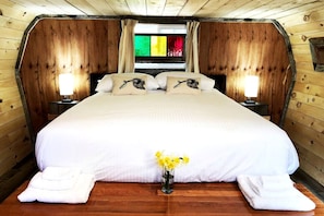 A wooden cabin-style bedroom with a large bed, two bedside lamps, and a window with colorful curtains. - Herons Haven Cabin in Nova Scotia (Baddeck)