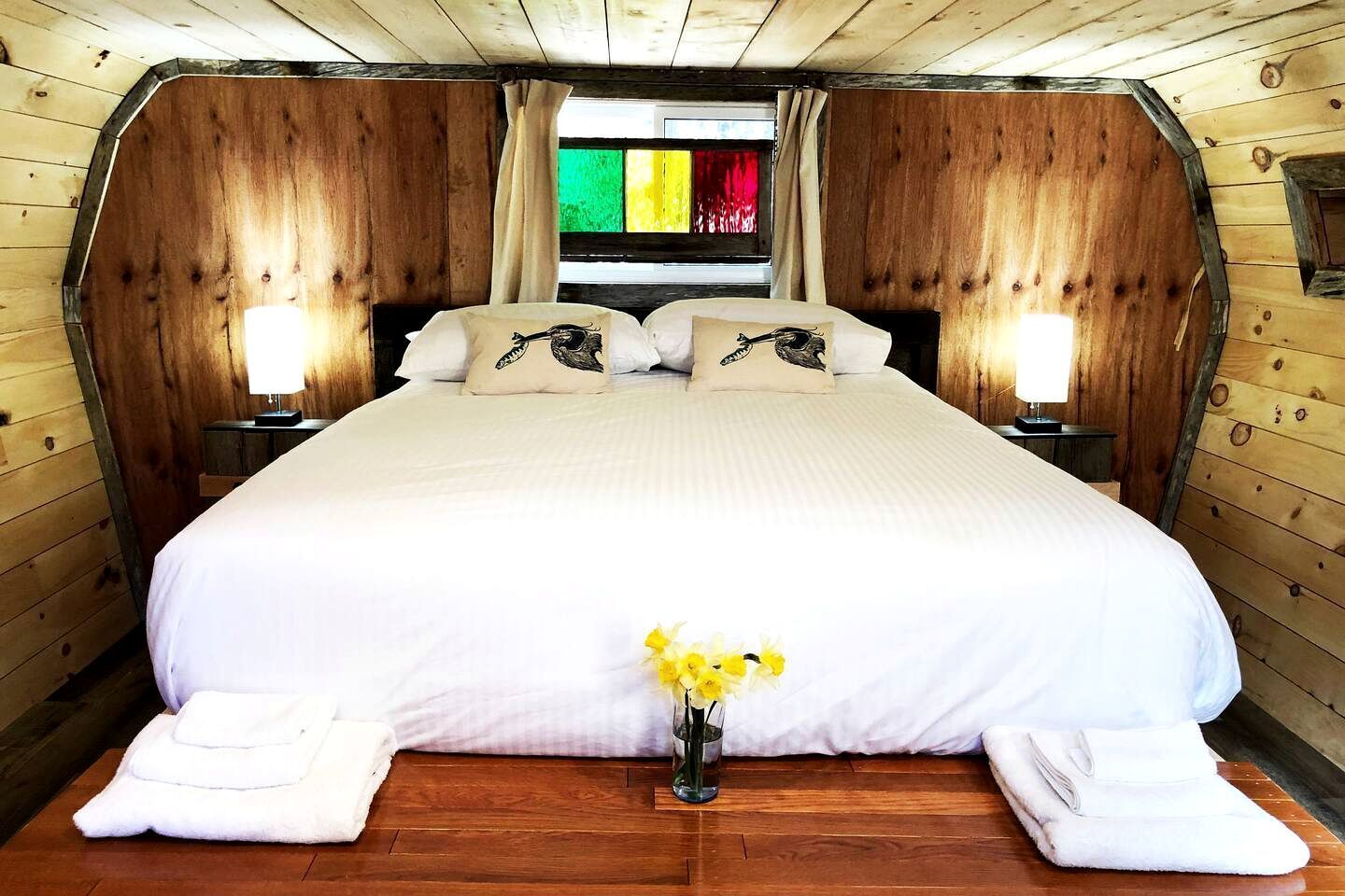 A wooden cabin-style bedroom with a large bed, two bedside lamps, and a window with colorful curtains.