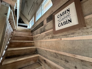 A wooden cabin interior with a staircase, wooden walls, and a sign that reads 'CABIN Sweet Cabin.'