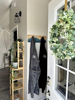 Ein Zimmer mit einem hölzernen Regal, auf dem Pflanzen stehen, einem schwarzen Robe an einem Haken und einem Fenster mit einem grünen Kranz.