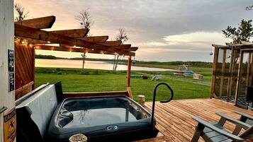 A hot tub on a wooden deck with a view of a lake and grassy fields.