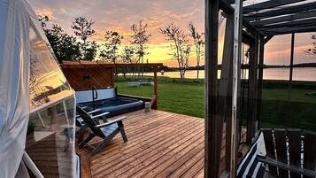 Eine Holzterrasse mit einem Zelt, einem Liegestuhl und einer Glaskonstruktion mit Blick auf einen Sonnenuntergang über dem Wasser.