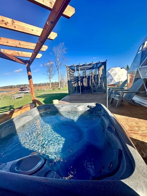 Ein Whirlpool mit einer hölzernen Pergola darüber, eine überdachte Sitzecke und Blick auf eine natürliche Landschaft.