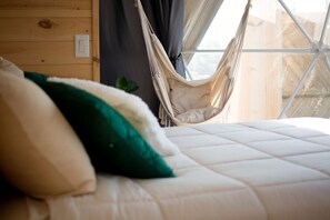 Ein Bett mit weißer Decke, einem grünen Kopfkissen und einer beigen Hängematte vor einer Holzwand mit einem Fenster.