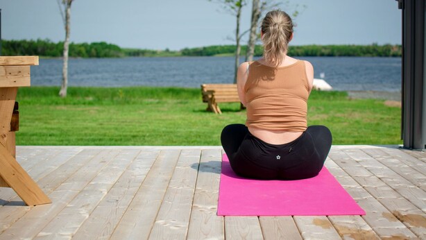 En person sidder på en lyserød yogamåtte på en træterrasse ved en sø, med en bænk og træer i baggrunden.