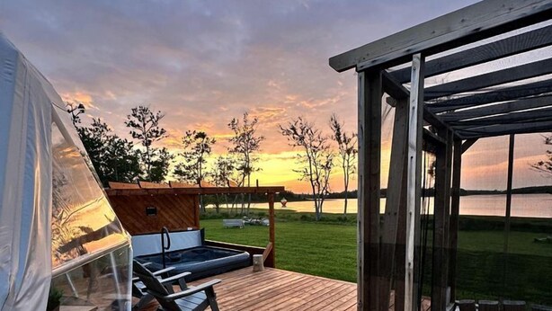 Une terrasse en bois avec une tente, un fauteuil de relaxation et une véranda fermée par des baies vitrées, offrant une vue sur un coucher de soleil au-dessus d’une étendue d’eau.