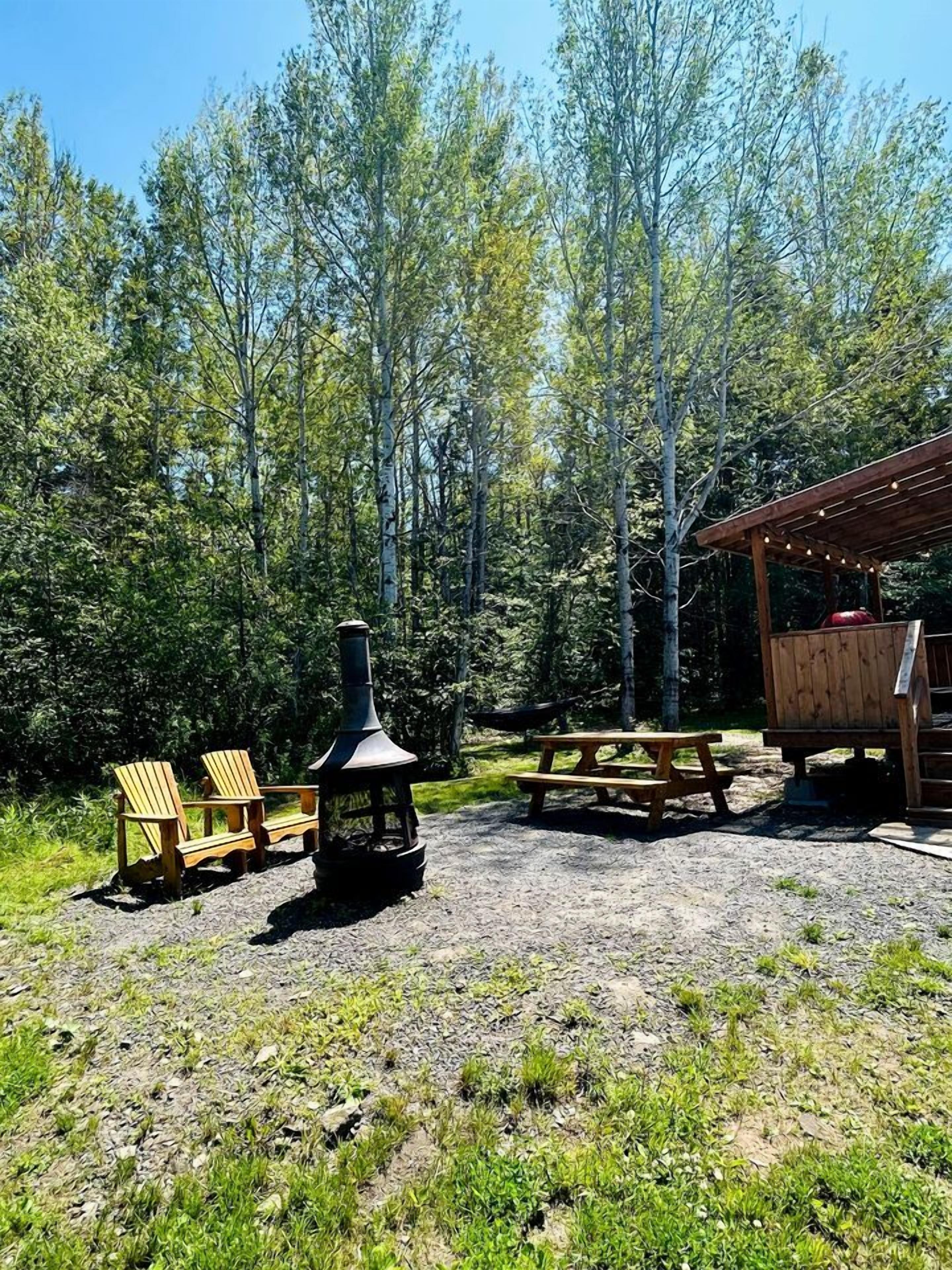 Eine rustikale Outdoor-Umgebung mit Adirondack-Stühlen, einem Holzofen, einem Picknicktisch und einem hölzernen Schutzdach.