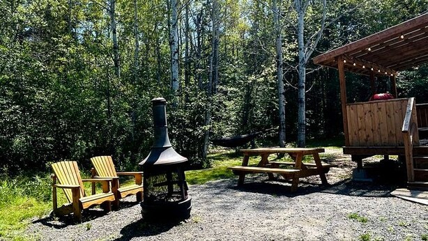 A rustic outdoor setting with Adirondack chairs, a wood-burning stove, a picnic table, and a wooden shelter. - Peaceful Boat Retreat with Luxury Interior with Hot Tub in Cocagne, New Brunswick (Cocagne)