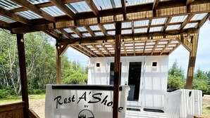 A wooden deck with a covered seating area, a small white building labeled 'Rest A' Shop,' and a natural outdoor setting. - Peaceful Boat Retreat with Luxury Interior with Hot Tub in Cocagne, New Brunswick (Cocagne)