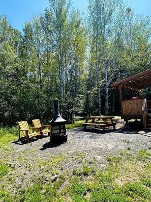 A rustic outdoor setting with Adirondack chairs, a wood-burning stove, a picnic table, and a wooden shelter.