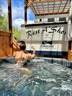 Une femme dans un bain à remous, avec une terrasse en bois et un panneau indiquant : « Espace détente A'Shoo, par CREENSIDE RNR ».