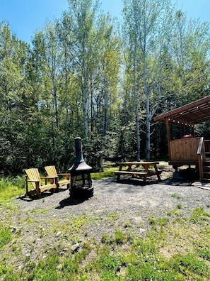 Un cadre extérieur rustique agrémenté de fauteuils Adirondack, d’un poêle à bois, d’une table de pique-nique et d’un abri en bois.