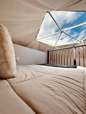Une pièce aménagée sous une tente, avec un lit, un oreiller moelleux, une barrière métallique et une vue sur le ciel.