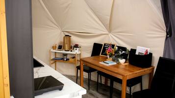 A tented room with a wooden table, chairs, a small shelf with items, and a welcome sign.
