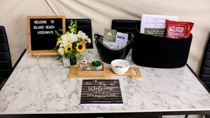 A marble table with a welcome sign, a guest book, a flower vase, a basket of snacks, and a thank you card.