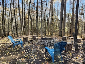 Un décor forestier avec un foyer, deux chaises Adirondack bleues et un banc en bois.