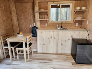 A wooden interior with a kitchen area including a table, chairs, a refrigerator, and a window with shelves above.