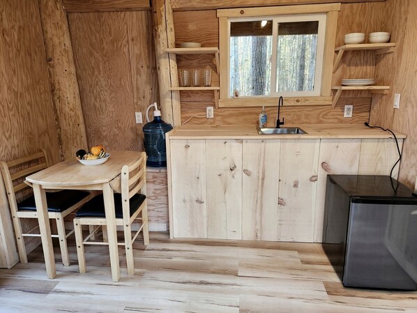 A wooden interior with a kitchen area including a table, chairs, a refrigerator, and a window with shelves above.