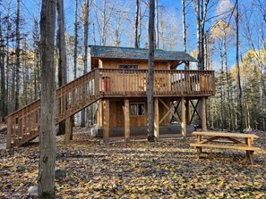 A wooden cabin with a balcony, a wooden deck, and a picnic table in a forest.
