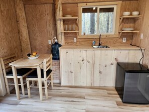 Puisesti sisustettu tila, jossa on keittiöalue. Keittiöalueeseen kuuluu pöytä, tuolit, jääkaappi ja ikkuna, jonka yläpuolella on hyllyjä.
