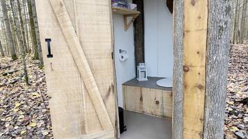 A wooden cabin with a door open, revealing a small bathroom with a toilet, sink, and shelf with toiletries.