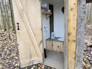 A wooden cabin with a door open, revealing a small bathroom with a toilet, sink, and shelf with toiletries.
